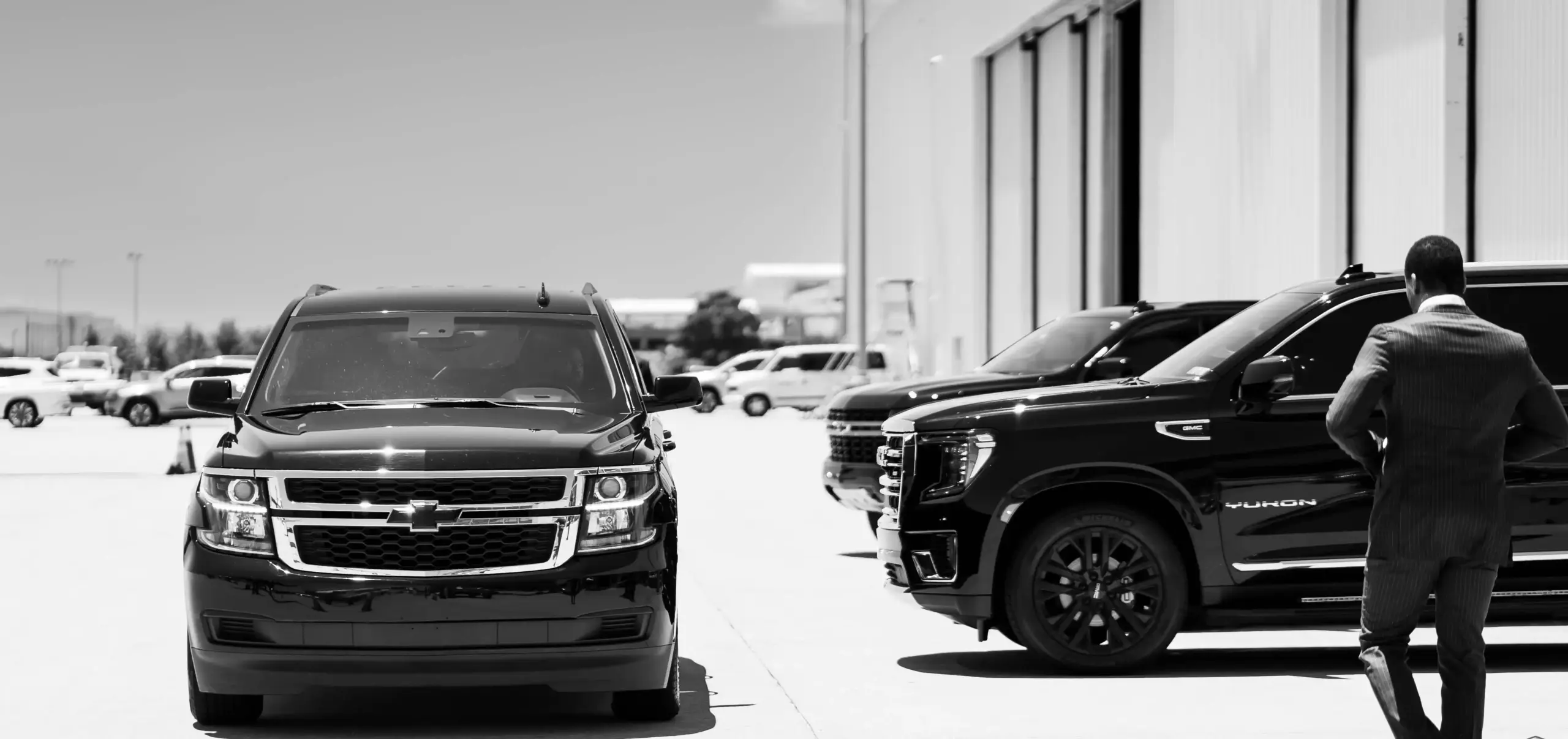chauffeur in dark suit checking tablet beside black executive SUV outside Boeing Field fixed-base operator, passenger boarding with rolling luggage and leather briefcase