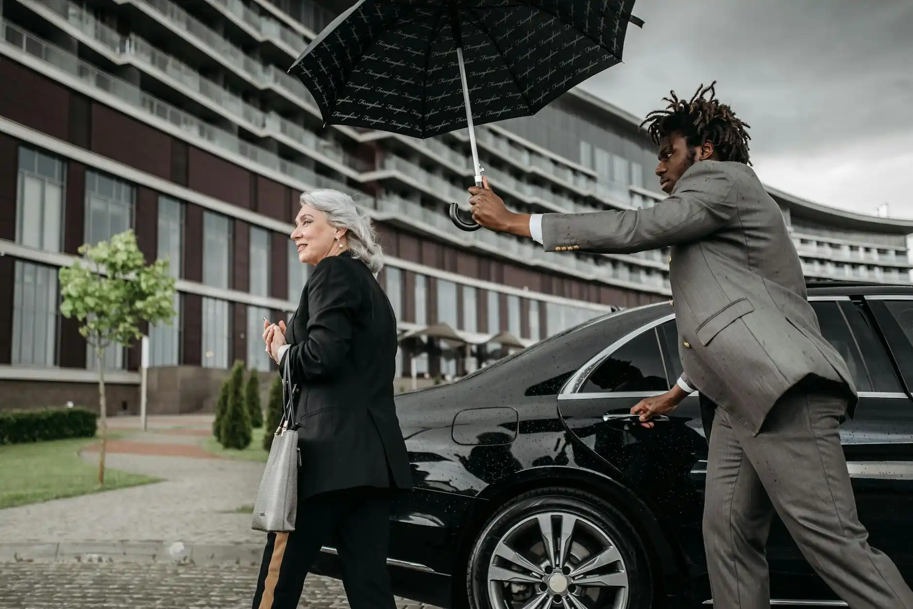 professional chauffeur standing beside a black executive limousine outside a downtown Seattle office tower, business travelers with briefcases entering vehicle, early morning ur...