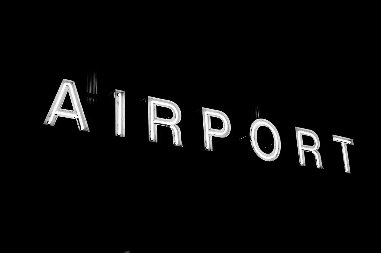 black luxury SUV curbside at Seattle-Tacoma International Airport at night, glowing terminal lights reflecting on wet pavement, traveler with rolling suitcase approaching driver...