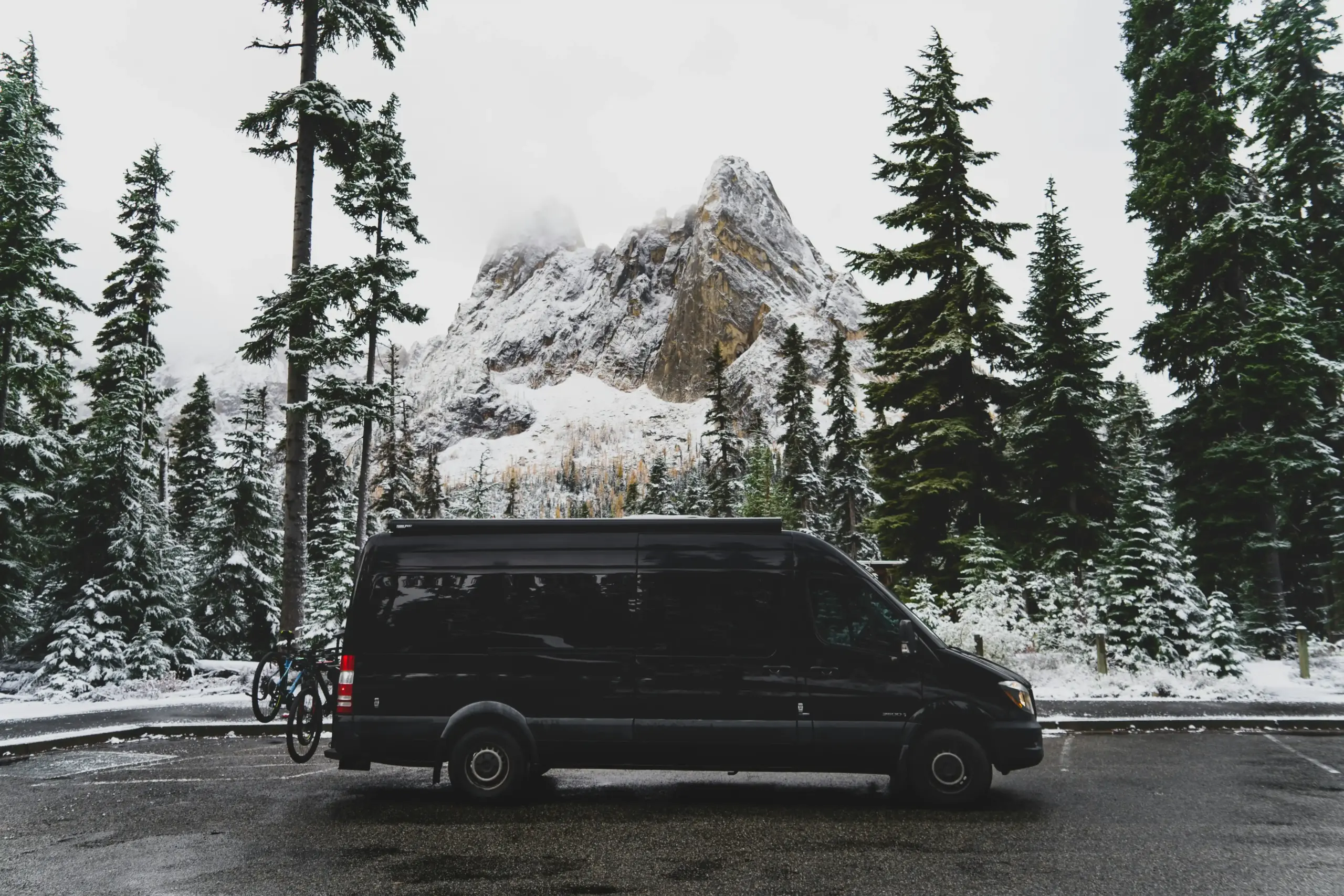 luxury black Sprinter van driving along a scenic Washington highway with evergreen forests, mountain backdrop, and soft morning light through low clouds